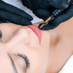 Young woman having permanent makeup on her lips at the beauticians salon. Natural green background